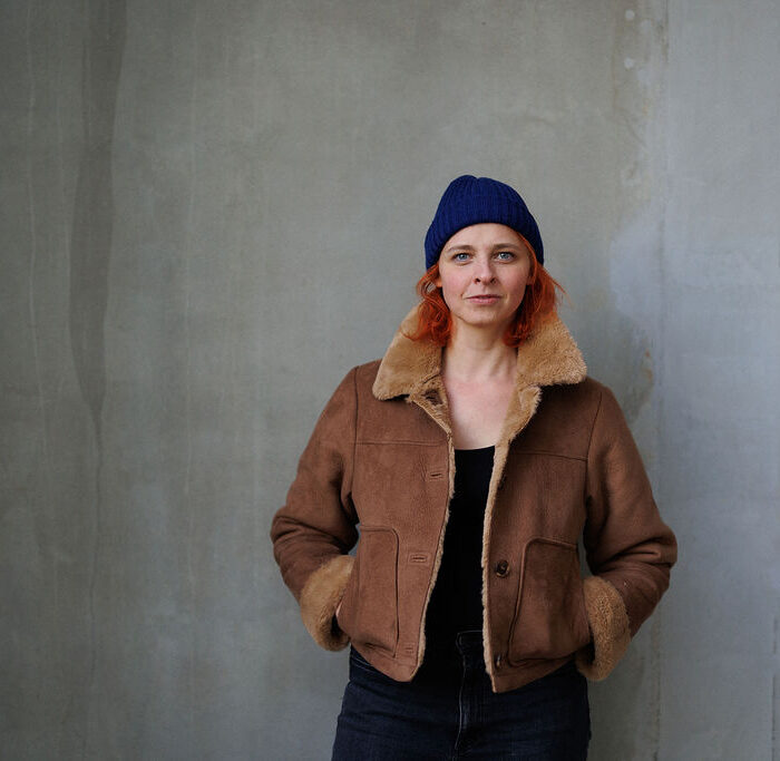 Autorin Annika Scheffel steht mit den Händen in den Jackentaschen vor einer grauen Wand und schaut in die Kamera